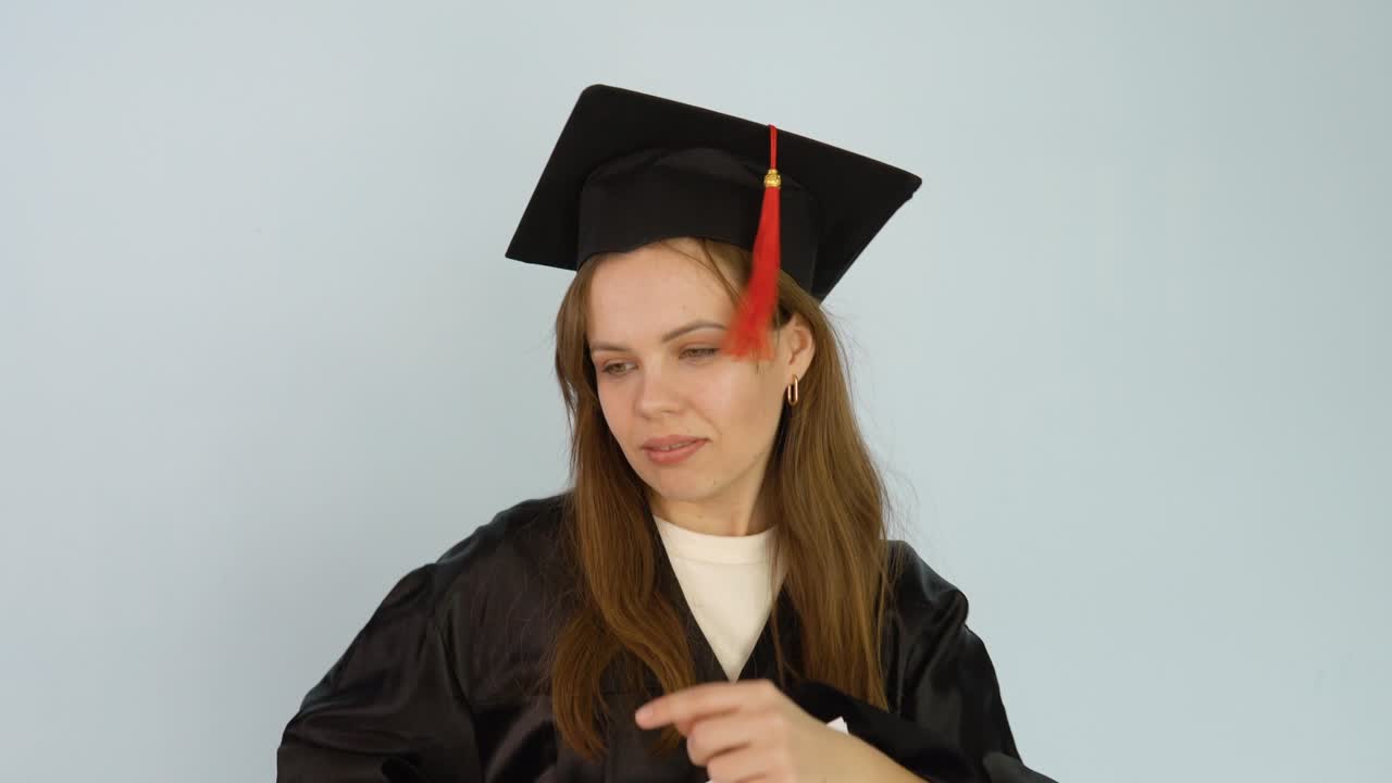 A university graduate dances with a diploma in her hands. Young female student in a black gown and a master's hat holds in an upright position a diploma of higher education. White background