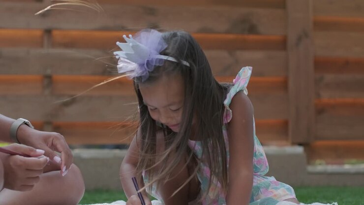 Una madre y su hija pequeña sentadas en una manta en el patio trasero, realizando una actividad de dibujo juntas. La escena al aire libre captura un momento familiar pacífico, fomentando la creatividad y el vínculo