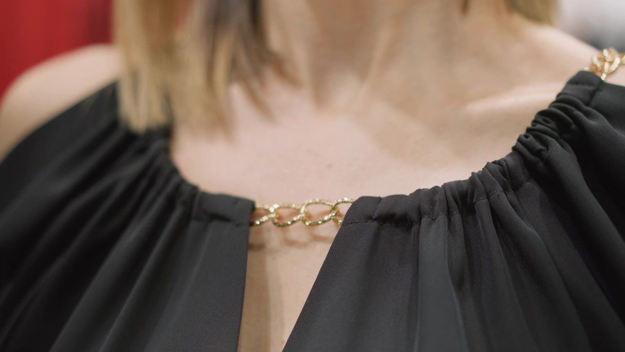 Primo piano di donna che indossa un top nero a spalle scoperte con dettaglio di catena d'oro intorno alla scollatura. La texture del tessuto e l'illuminazione soffusa evidenziano le proprietà del materiale del capo su sfondo rosso sfocato.