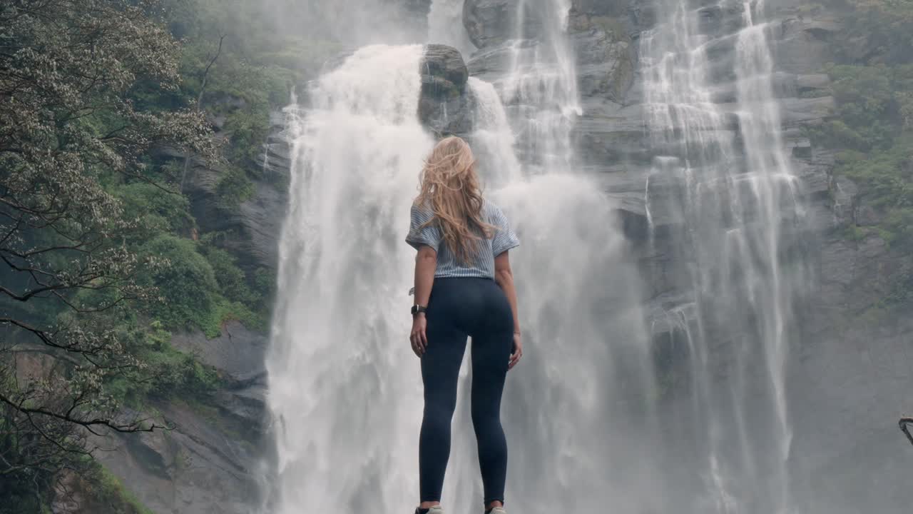 A woman stands in awe before the roaring Bomburu Ella Waterfalls, surrounded by mist, jungle, and rocky cliffs in the lush highlands of Nuwara Eliya, Sri Lanka.