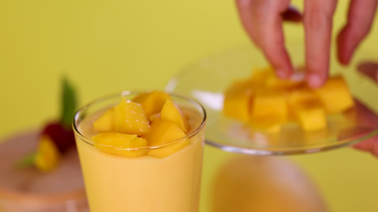 Hand adds mango cubes to smoothie in glass, bright yellow background, soft natural lighting, close-up