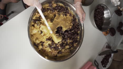 overhead shot of cookie dough with chocolate cubes being mixed with spatula in a stainless mixing bowl