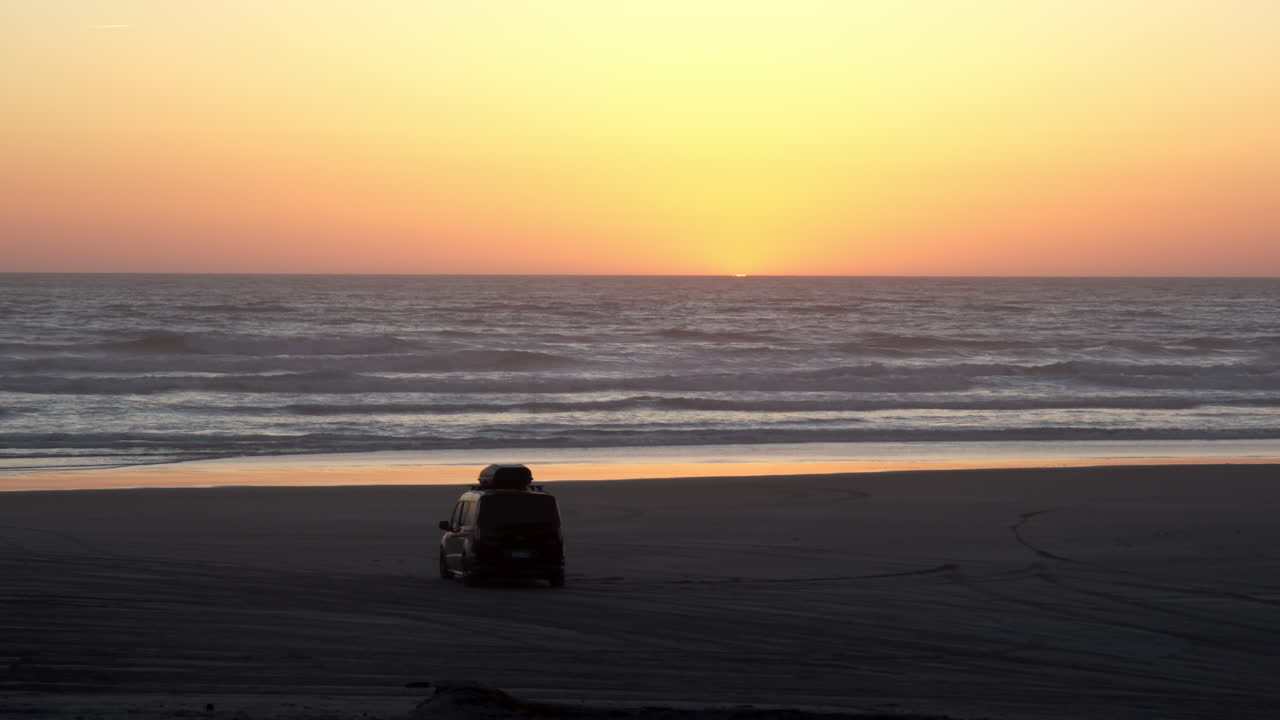 A lone SUV sits on a quiet beach at sunset as waves roll in beneath a glowing orange horizon
