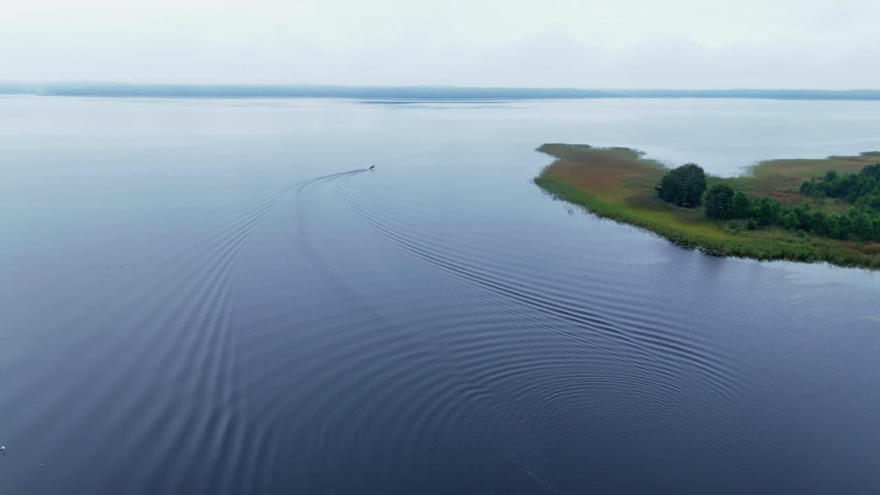 Drone shot captures small boat leaving visible wake across open blue water surrounded by marsh vegetation and forested shoreline on Usma Lake in Kurzeme region