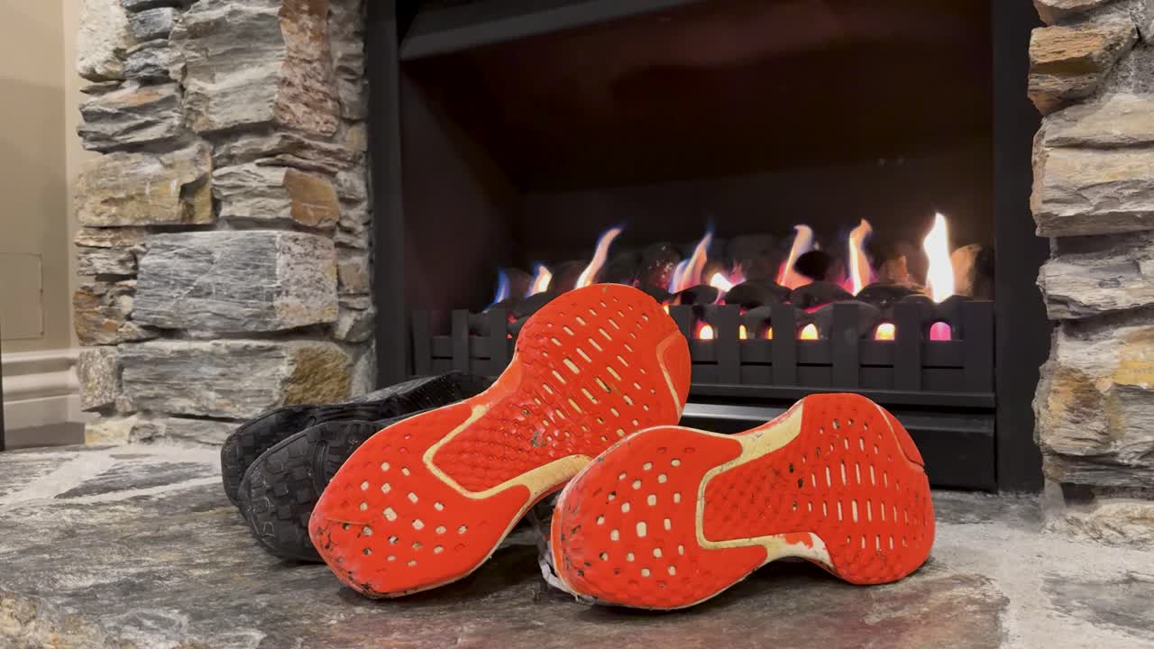 A pair of orange-soled shoes rests on stone flooring in front of a lit fireplace, with warm ambient lighting and a rustic interior setting