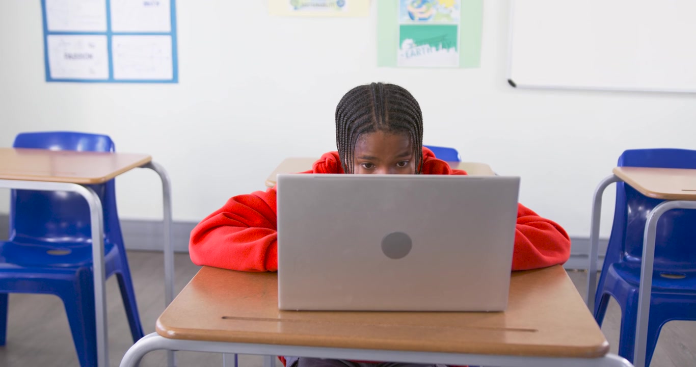 Adolescente afroamericano che lavora al laptop al banco di scuola completando un compito mentre la telecamera si allontana.