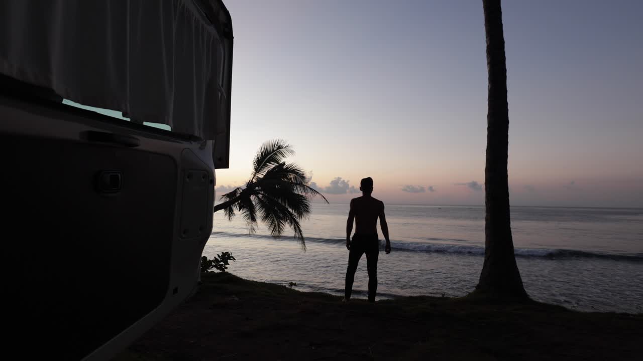 joven caminando al borde de una playa durante el amanecer con vistas a los cocoteros con la puerta de la caravana abierta en bali, indonesia