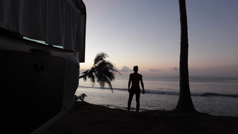 young man walking to the edge of a beach during sunrise overlooking the coconut trees with the camper van door open in bali indonesia