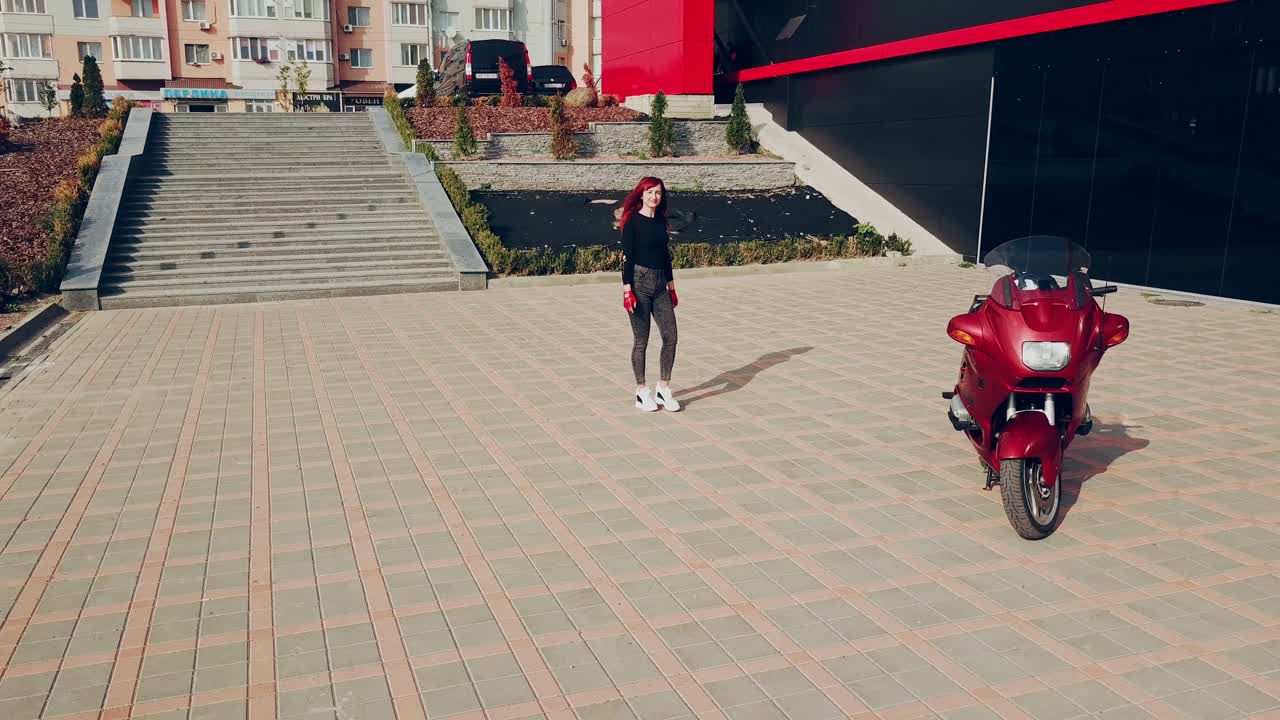 Young sexy woman wearing grey jeans and black shirt with red gloves goes to the new motorcycle. Happy female sitting on a red motorbike.