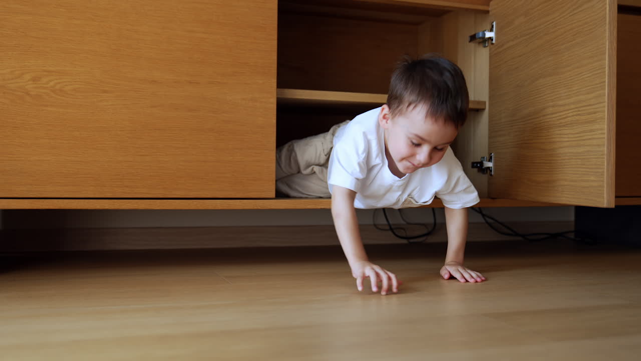 Baby boy lies inside the wooden nightstand. Happy toddler climbs out of the chest and smiles to camera