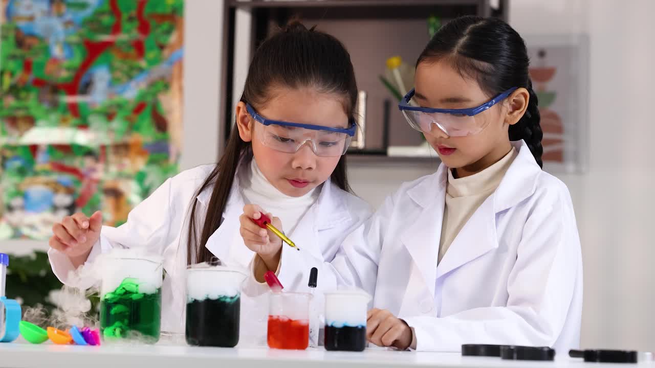 Mahasiswa Asia mengenakan jas lab menggunakan pipet untuk percobaan kimia berwarna-warni di laboratorium yang terang.