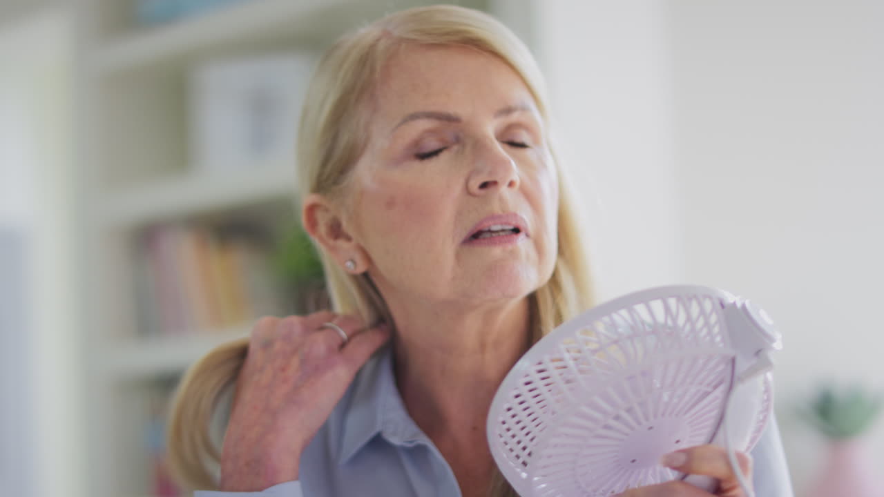 mujer madura en la menopausia que tiene sofocos en casa refrescándose con un ventilador eléctrico