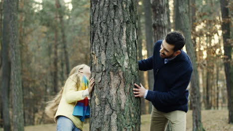 joyful caucasian father and daughter playing in the wood, hiding behind a tree trunk and looking at each other from different sides