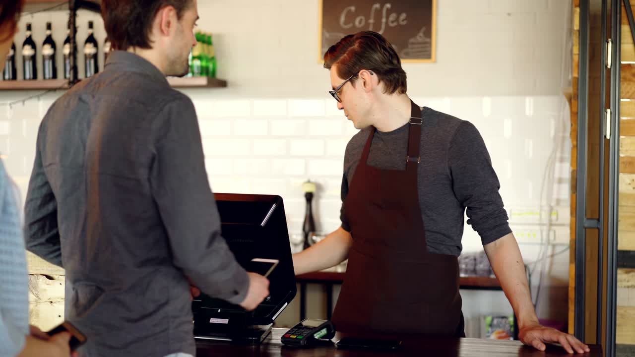 Friendly barista handsome man is selling takeout coffee and accepting contactless mobile payment with smart phone. Modern technolofy, coffee shop and banking concept.