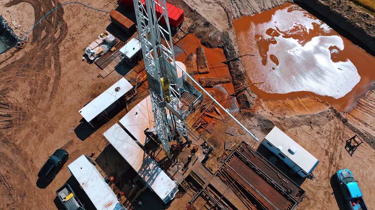 Rising along the rig for drilling oil. Few men are working at the site. Lots of metal pipes are next to the derrick. Water flows to the pool with orange dirt.