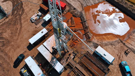 rising along the rig for drilling oil. few men are working at the site. lots of metal pipes are next to the derrick. water flows to the pool with orange dirt.