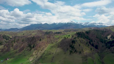 Vista-Aérea-De-Una-Montaña-Cubierta-De-Nieve-Con-Un-Cielo-Azul-Claro,-Nubes-Blancas-Y-Vegetación-Verde-Con-Una-Pequeña-Casa-En-La-Cima-De-Una-Colina,-Rumania