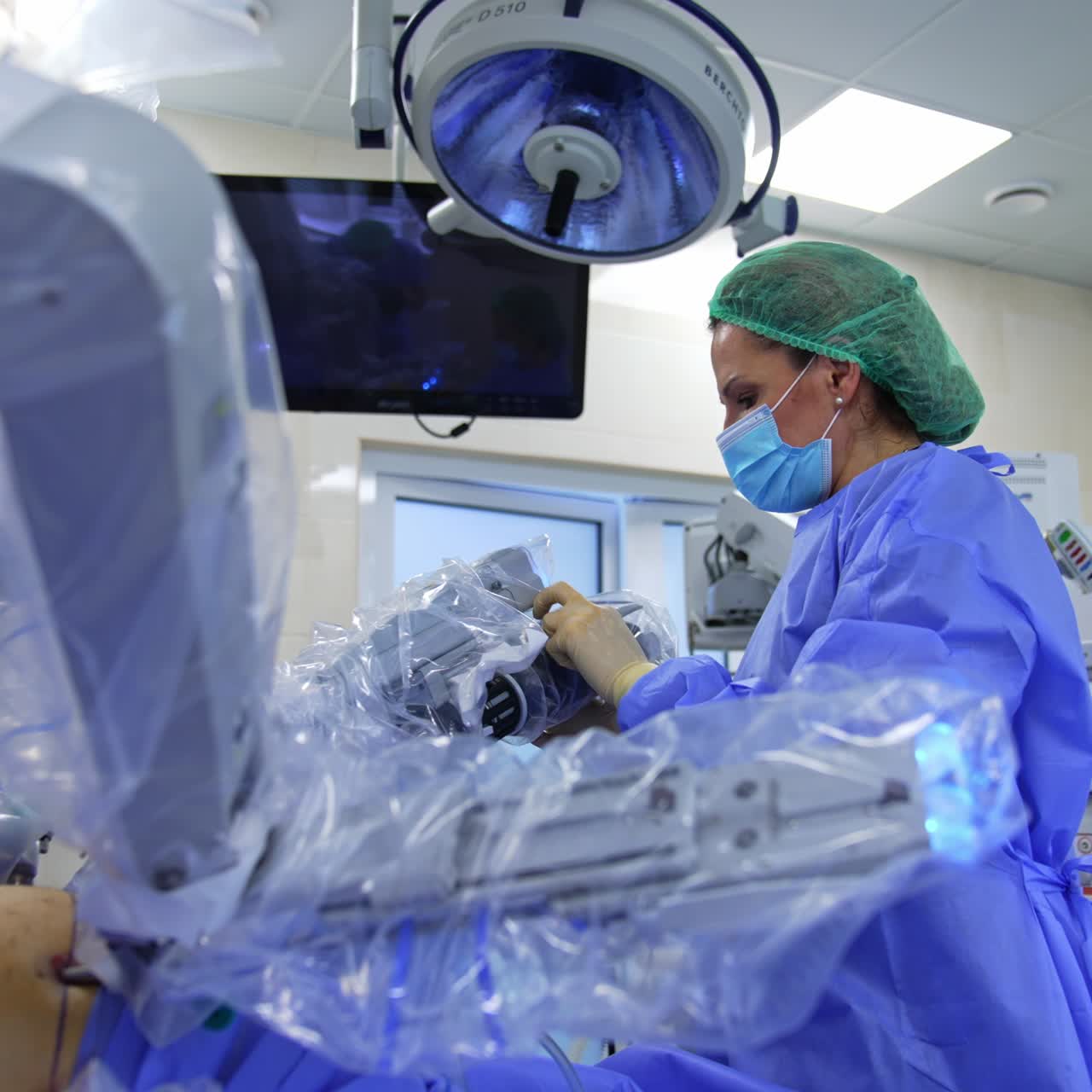 Woman in mask, cap and uniform working in the surgery room. Medic touches the robotic arms operating the patient and looks at the screen