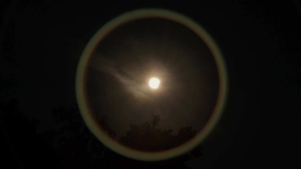 Start of the Solar Eclipse of 25.07.2026 the sun is slowly moving in front of the sun making a circular lens reflection on the lens just above some tree branches and clouds.