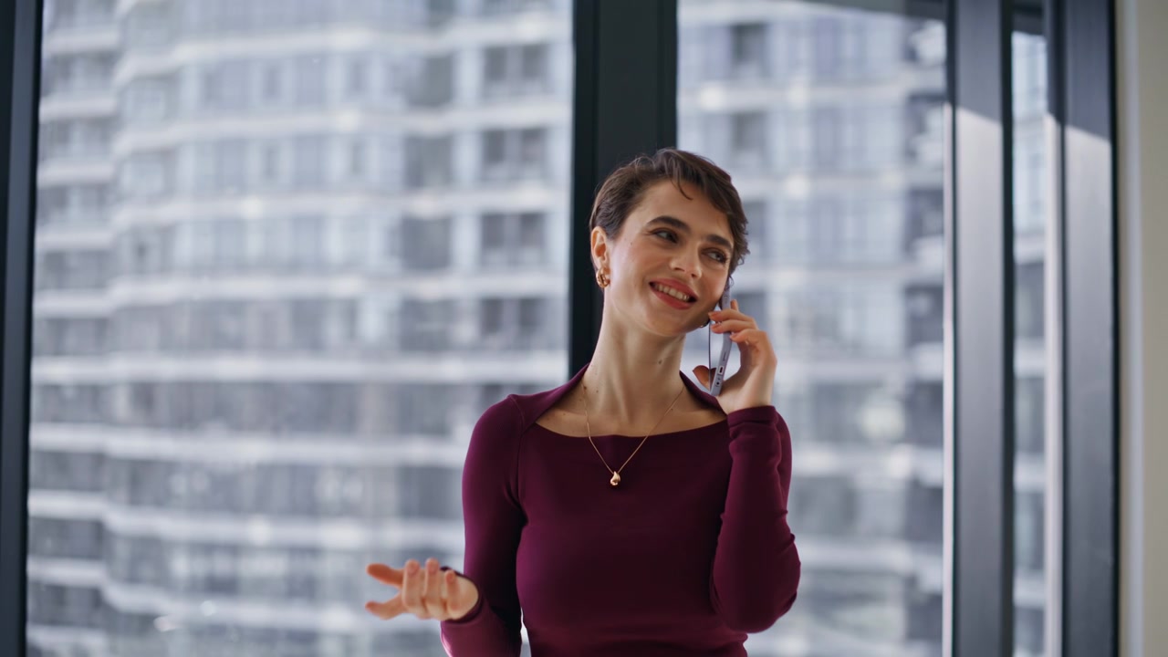 Glad kvinne snakker i smarttelefon ved kontorvinduet. Smilende dame i telefonsamtale innendørs.