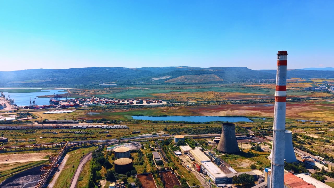 Vista dall'alto di un impianto industriale costiero e di un porto container. Una ciminiera rossa e bianca si erge vicino a torri di raffreddamento, accanto a banchine portuali e campi verdi.
