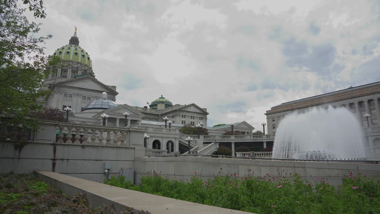 Panorâmica cinematográfica para a direita através da fonte no Capitólio Estadual da Pensilvânia, em Harrisburg, Pensilvânia, revelando arquitetura governamental histórica e um proeminente marco cívico.
