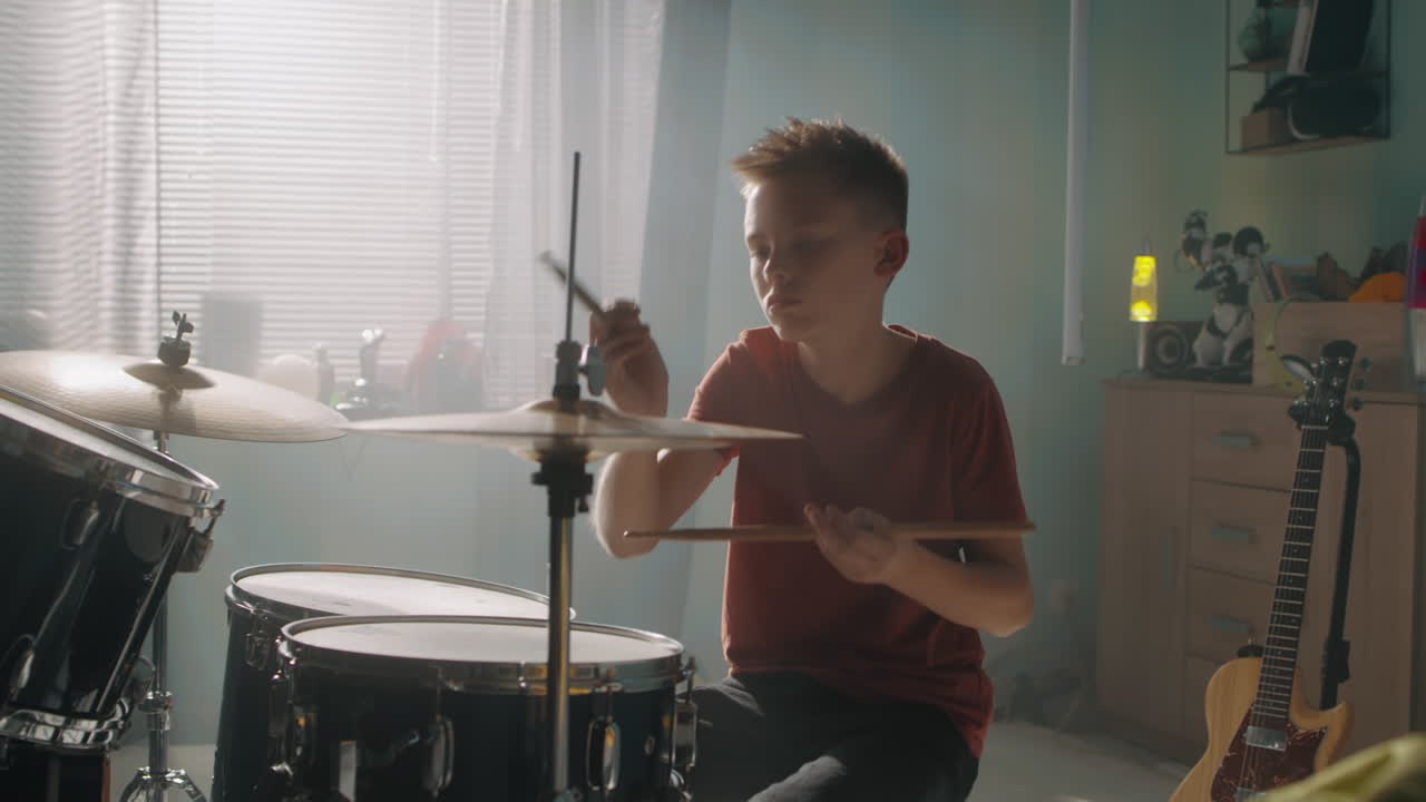 Teen Drummer Rehearsing in Bedroom Teenager in Casual Clothes Sitting Near Bed and Playing Drums during Rehearsal in Illuminated Bedroom at Home
