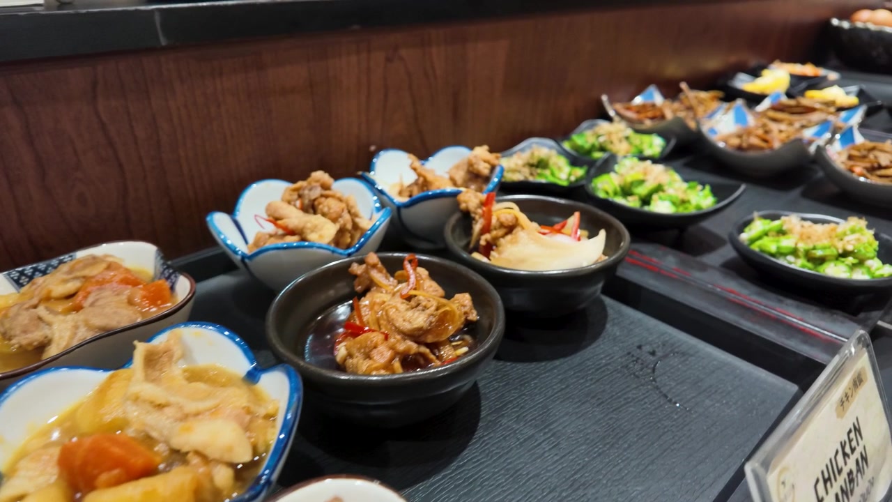 Kameraet panorerer hen over forskellige keramikskåle med japanske sideretter på en buffetrestaurant i Bangkok.