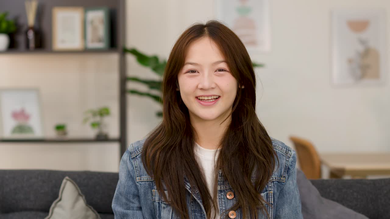 หญิงสาวร่าเริงบนโซฟายิ้มให้กล้อง พร้อมทำท่าโอเคในห้องนั่งเล่นที่สว่างสดใส