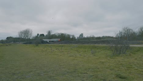 Long-shot-of-a-train-passing-by-a-grassy-field-in-the-countryside-on-an-overcast-day-in-late-fall-with-leafless-trees