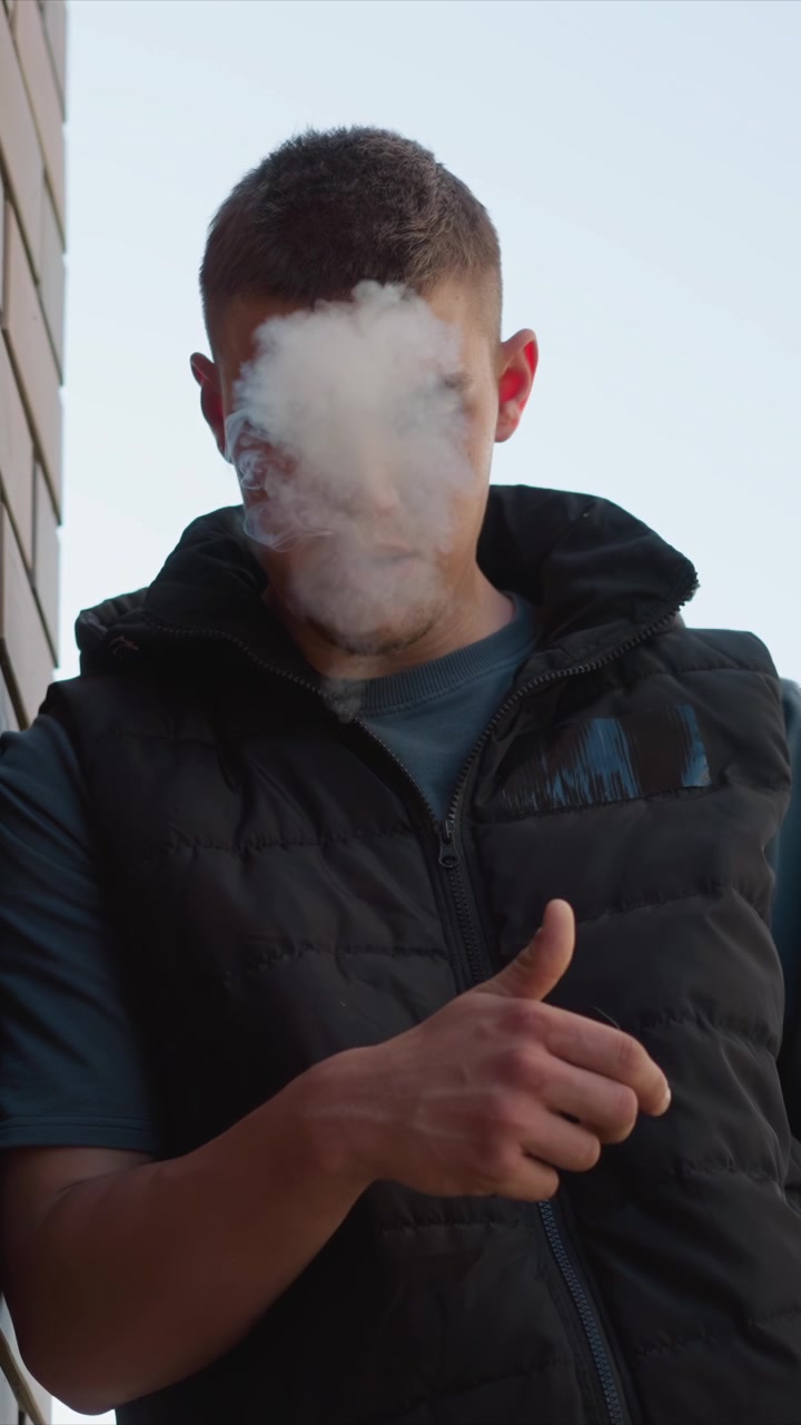 Primo piano di giovane uomo con gilet imbottito nero e camicia blu che esala fumo mentre è in piedi all'aperto accanto a un edificio con sfondo di cielo sereno. Gesti delle mani inclusi indicare e toccarsi il viso.