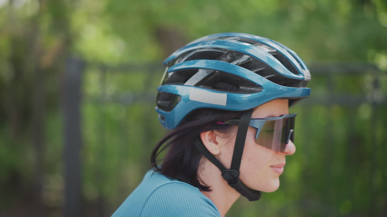 side profile woman wearing helmet, reflective sunglasses and calm expression in urban park closeup on teal helmet vents and chin strap, steady posture suggesting training ride or commuter routine