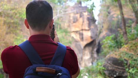 30s mountain hiking photographer or video blogger man in woods filming a canyon on cell phone. guy traveler in red polo and with backpack videographer standing in nature. back view.