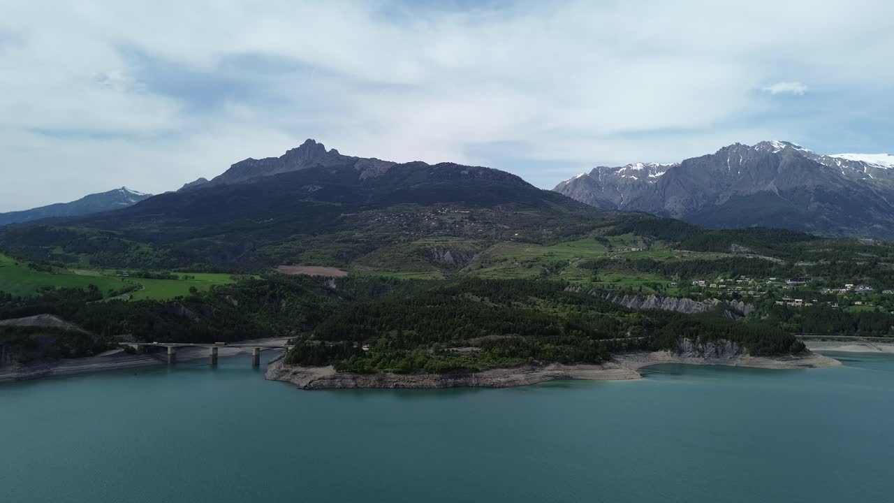 4K drone footage flying backward over Lac de Serre Ponçon, showcasing the Aiguilles de Chabrières, Parc des Écrins, a bridge, lush green trees, and snowy mountain tops. Captured May 2025, French Alps