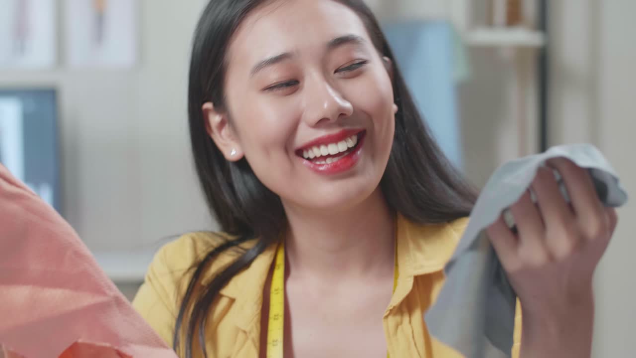 미소 짓는 아시아 여성 디자이너가 스튜디오에서 옷을 디자인하는 동안 직물을 들고 쳐다보고 있습니다.