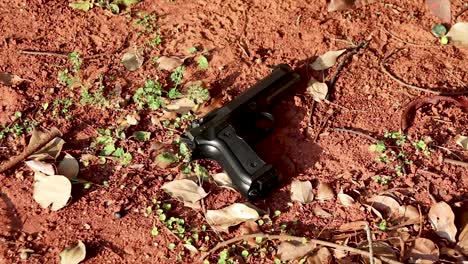 zooming in of a black firearm laying on a sandy ground with some dry scattered leaves