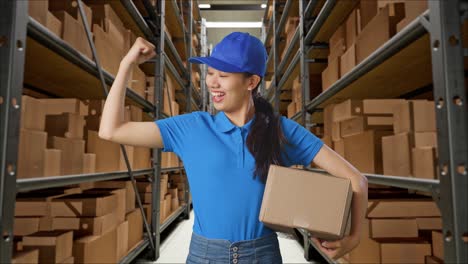 mensajera asiática en uniforme azul sonríe y flexiona los músculos mientras entrega una caja en el almacén