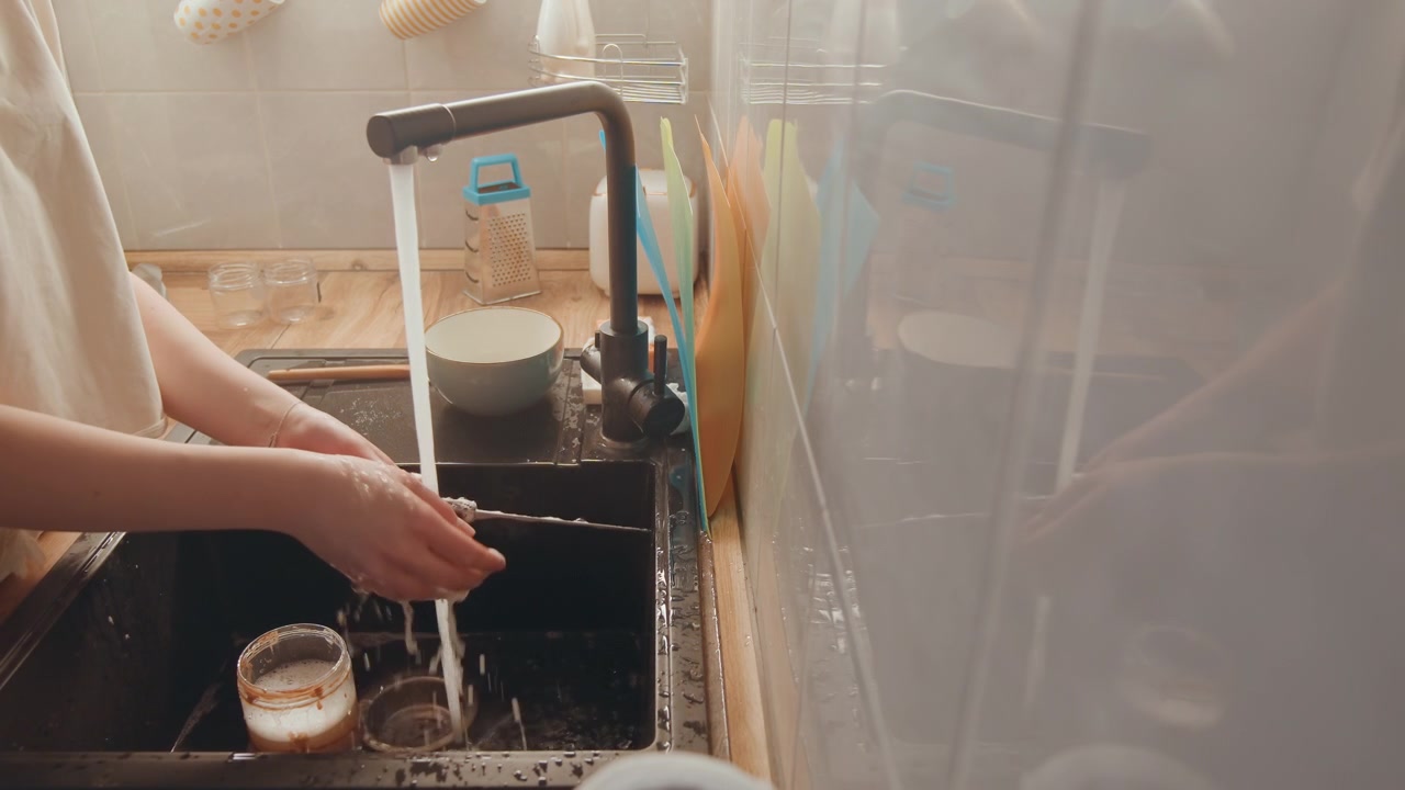 Kvinde vasker op. Kvinde rengør pande med vandstråle. Køkkenopgave med vask af pande med sæbeskum. Kvinde udfører opvask i et varmt, oplyst køkkenmiljø.