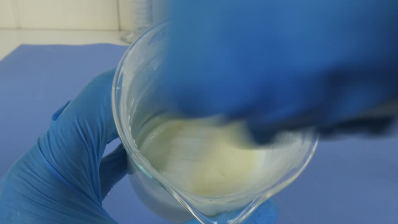 Macro shot of a cosmetic emulsion being prepared in a laboratory. The video captures the operator's gloved hands mixing ingredients in a beaker,