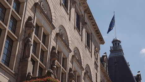 Toma-Panorámica-Del-Ayuntamiento-De-Rotterdam,-Edificio-De-Piedra-Arenisca-Con-Estatuas-Sobre-La-Entrada-Con-Una-Bandera-Europea-Ondeando