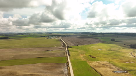 Eine-Luftaufnahme-Zeigt-Eine-Weitläufige-Landschaft-Mit-Landwirtschaftlichen-Feldern-In-Polen,-Mit-Unterschiedlichen-Schattierungen-Von-Grün-Und-Braun-Unter-Einem-Dramatischen,-Mit-Wolken-übersäten-Himmel,-Einer-Straße,-Die-Sie-Durchschneidet