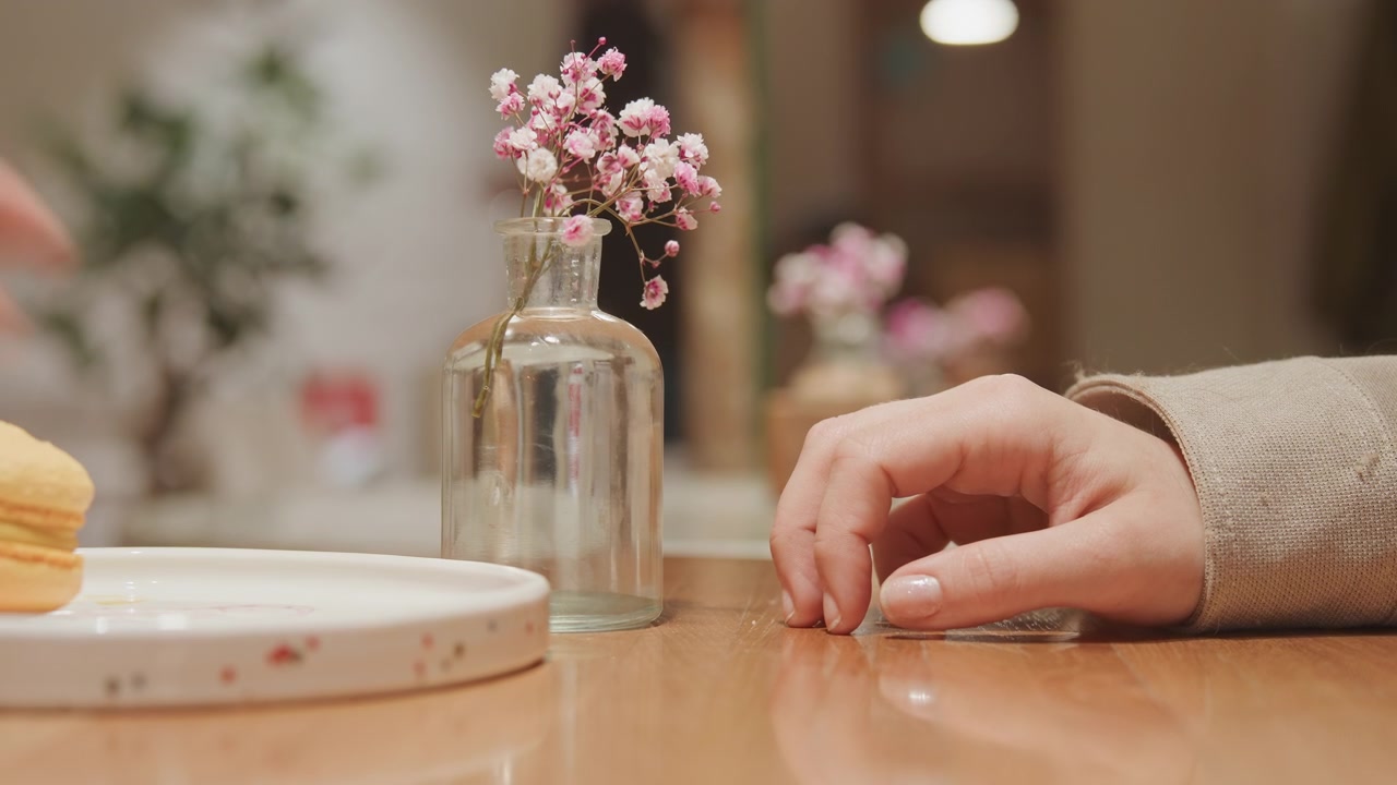 Gros plan d'une main posée sur une table en bois près d'un vase en verre transparent avec des fleurs roses, éclairage doux et arrière-plan flou. La main bouge légèrement tandis qu'une assiette est placée à côté du vase, maintenant la mise au point.