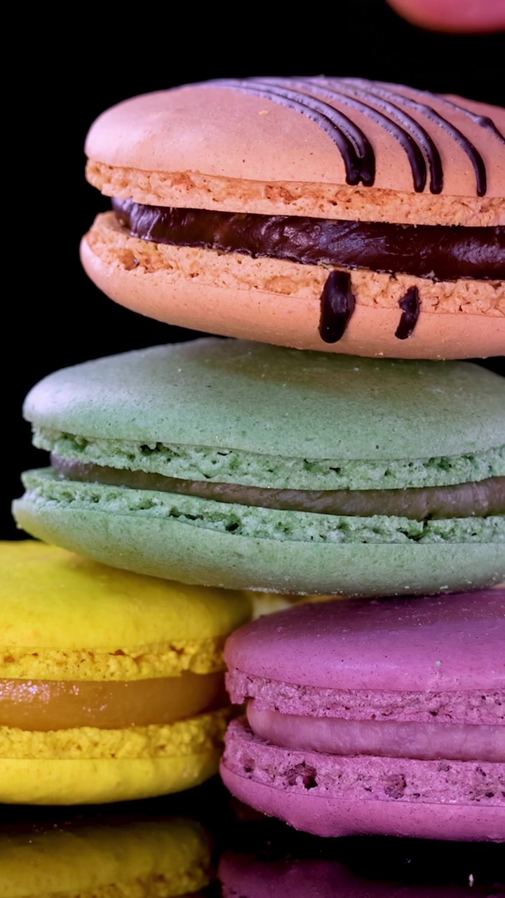 A hand carefully picks up and stacks assorted pastel macarons on a glossy black surface under bright, even lighting, emphasizing vibrant colors and dessert textures