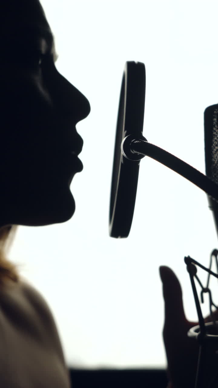 Silhouette of beautiful woman is singing a song in front of a microphone in an audio recording studio on a white background. Close-up Vertical video