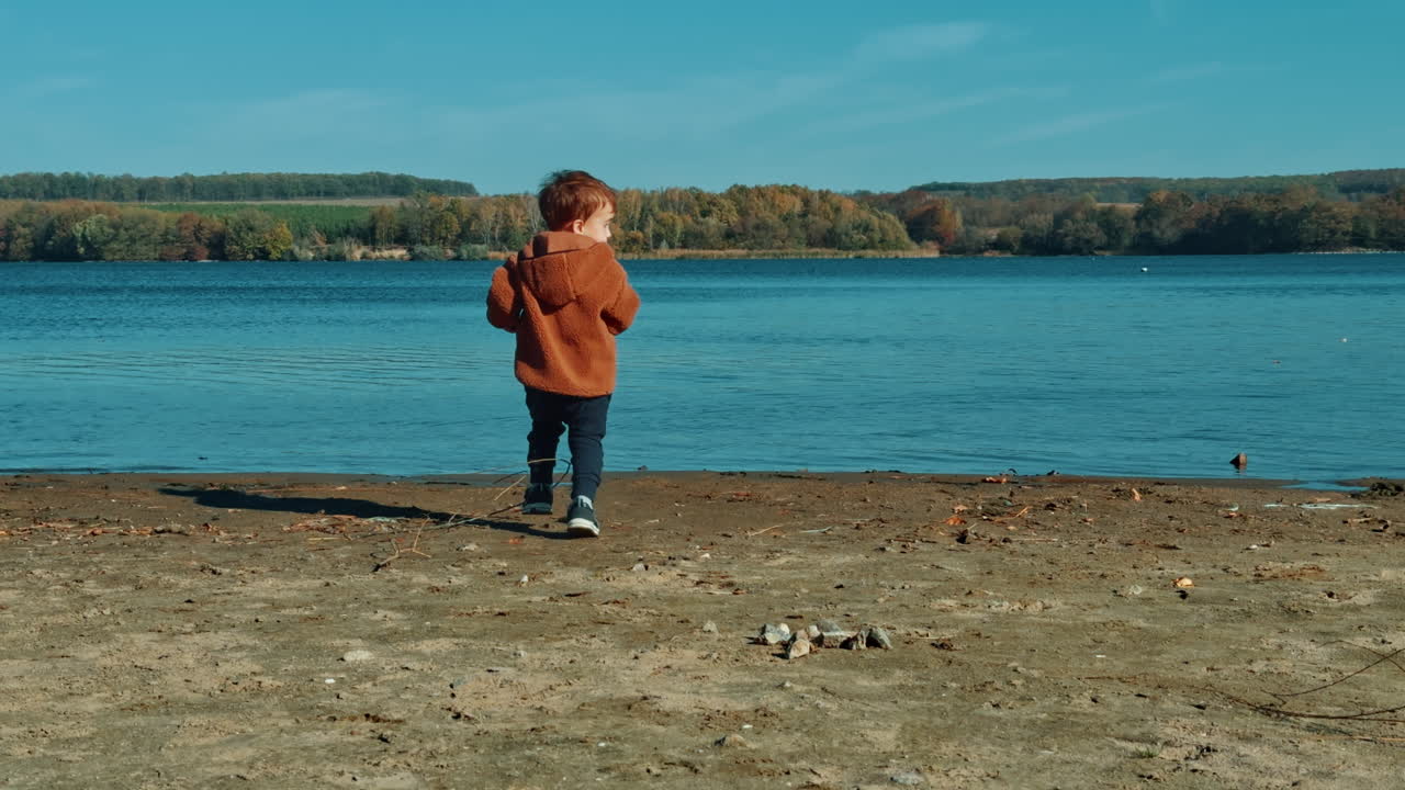 Energetic baby boy picking stones on the river bank. Kid in warm jacket throws stones into water.