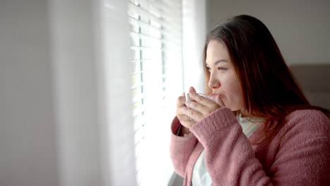 plus size biracial woman drinking tea and looking out of bedroom window, copy space, slow motion