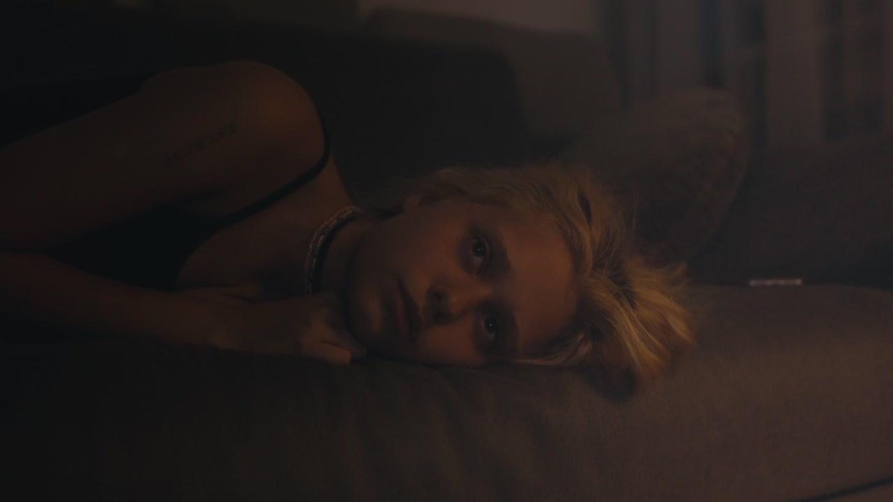 una joven rubia con una camiseta gris se acuesta y mira el techo durante el insomnio en el sofá en una habitación oscura por la noche