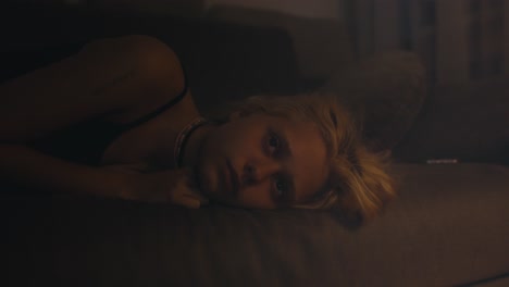 zoom in young blonde girl in a gray top lies and looks at the ceiling during insomnia on the sofa in a dark room at night