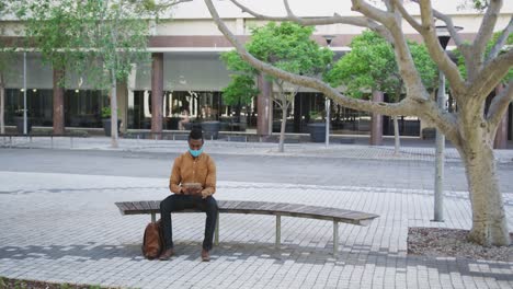 african american businessman wearing face mask using tablet sitting on bench