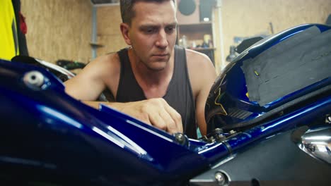 toma de primer plano: un hombre con una camiseta gris, un mecánico con un corte de pelo corto, está reparando su motocicleta gris en su taller - un garaje. inspección de equipos en el taller